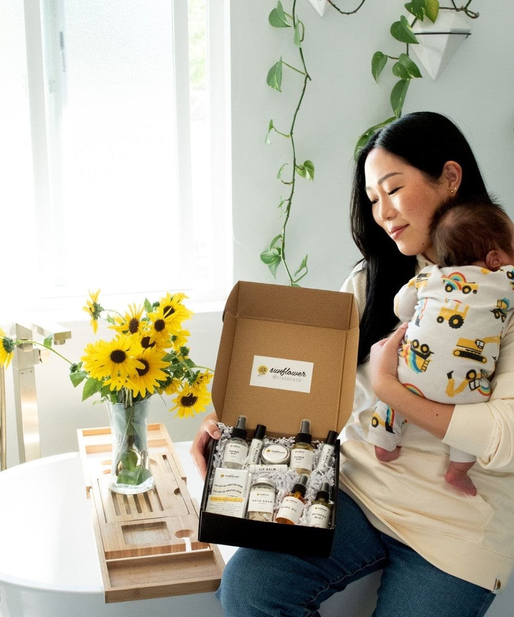 A woman holding a baby and a box of products with sunflowers and a wooden crate in the background.