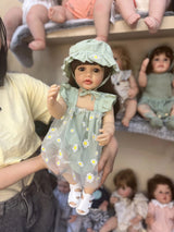 Doll in a green dress with floral patterns held by a person, surrounded by other dolls.