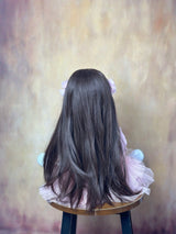 Back view of a doll with long dark hair sitting on a stool against a textured wall.