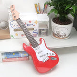 Red toy guitar on a white surface with stationery and a plant in the background