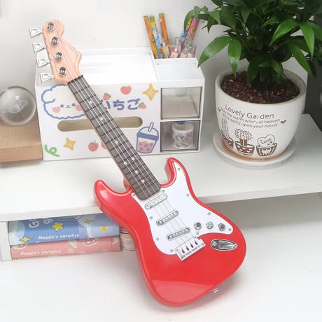 Red toy guitar on a white surface with stationery items and a plant in the background