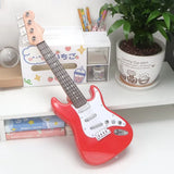 Red toy guitar on a white surface with a plant and stationery items in the background