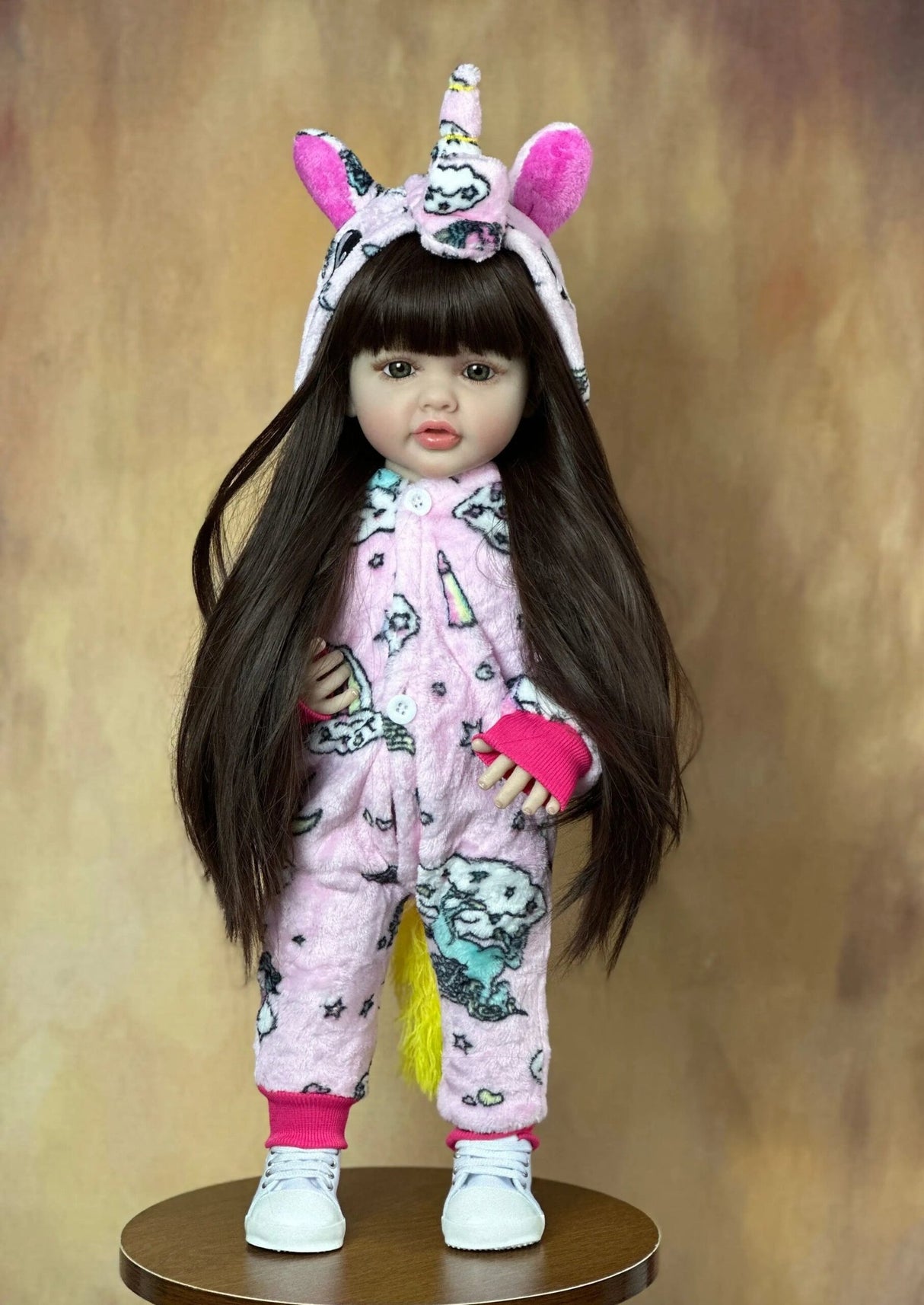 Doll wearing a pink unicorn onesie with a headband on a brown background