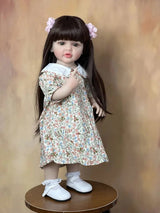 Doll wearing a floral dress and white shoes on a wooden pedestal against a brown background