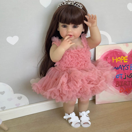 Doll in a pink dress with a tiara, standing against a decorative wall with hearts and a birthday card.