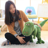 Woman sitting on the floor with a large green dinosaur plush toy in a bright room.