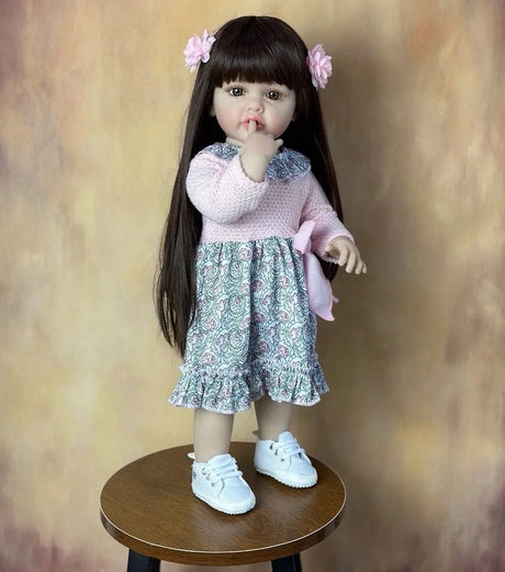 Doll with long dark hair wearing a pink sweater and patterned dress on a wooden stool