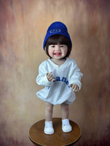 Child wearing a blue hat and white outfit with text, standing on a wooden platform