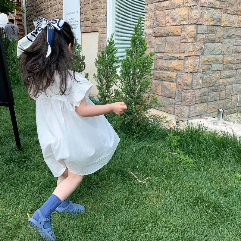 Child in a white dress and blue shoes standing on grass next to a stone wall.