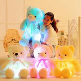 Woman holding a glowing teddy bear with three other colorful teddy bears on a bed.