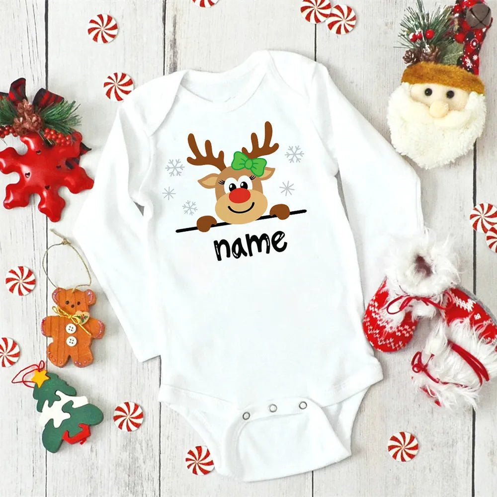 White baby onesie with a reindeer design and 'name' text, surrounded by Christmas decorations on a wooden surface. (option 23)