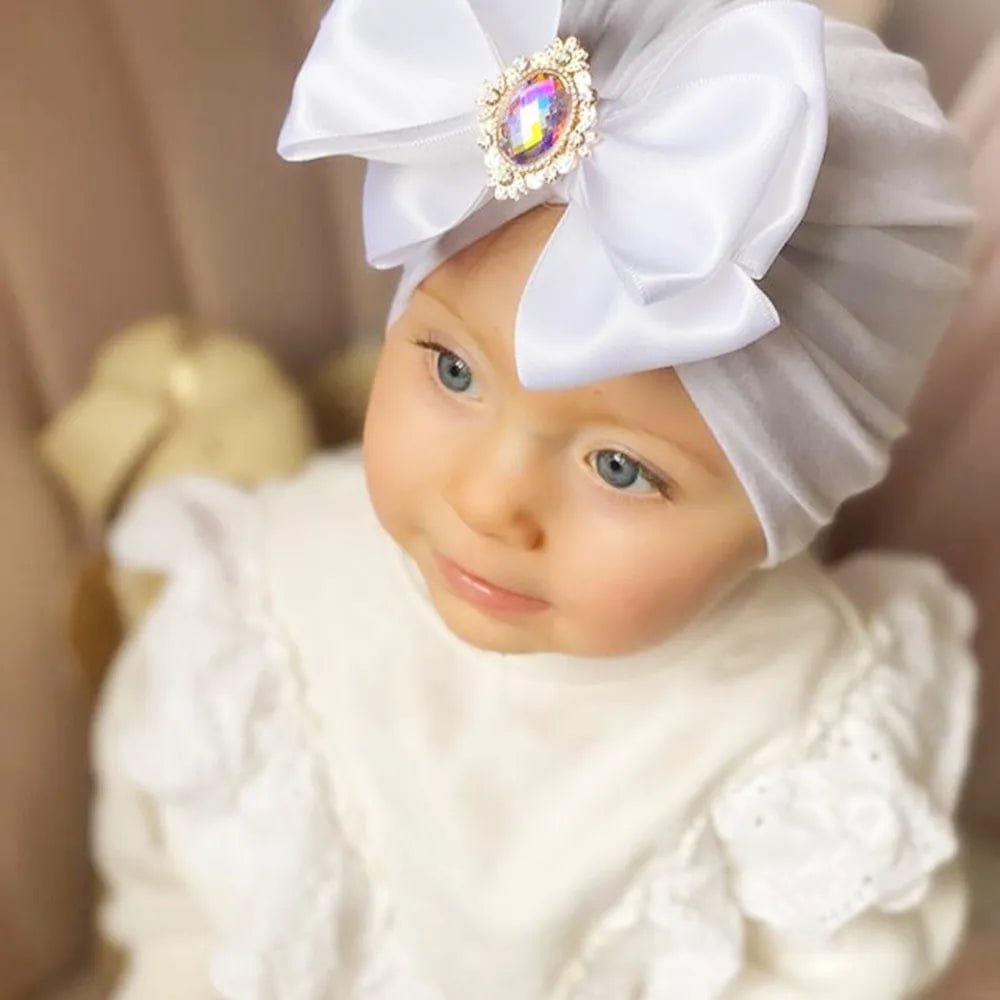 Baby wearing the white rhinestone bow turban cotton cap for gentle head coverage and dressed-up outings.