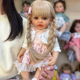 Person holding a doll with blonde hair and a pink dress, surrounded by other dolls in the background.