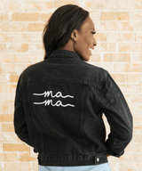 Person wearing a black denim jacket with 'ma ma' text on the back against a beige brick wall.