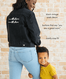 Woman wearing a black denim jacket with 'ma ma' text, standing next to a child against a brick wall.