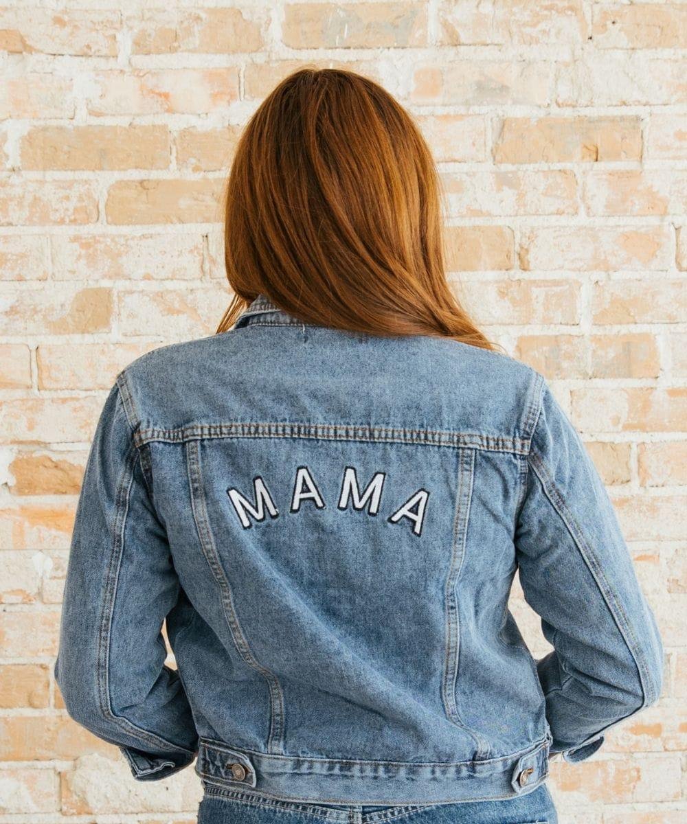 Person wearing a denim jacket with 'MAMA' on the back against a brick wall.
