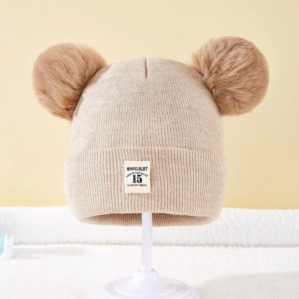Beige knit baby hat with two fluffy brown pom-poms on a light yellow background