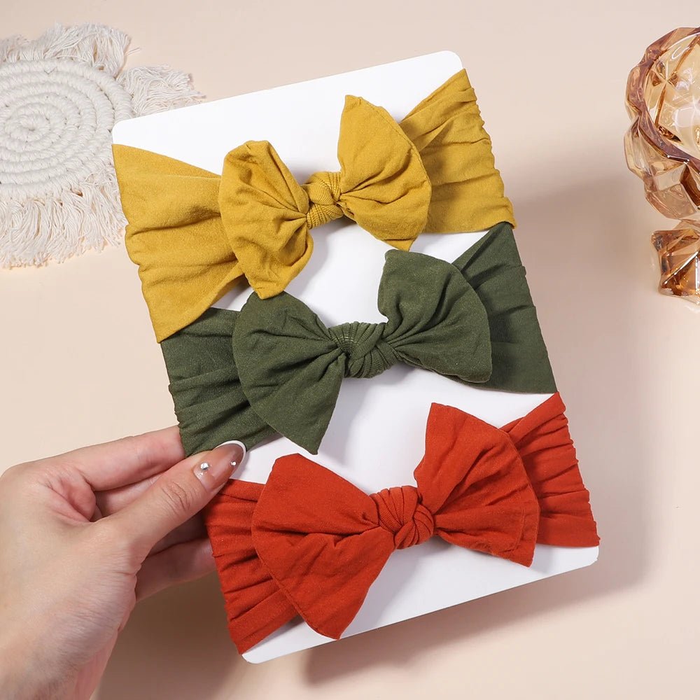Three fabric hair bows in yellow, green, and red on a white tray held by a hand.