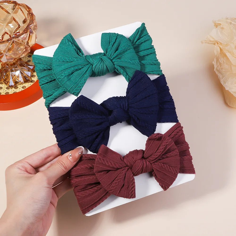 Three fabric hair bows in green, navy, and burgundy on a white box with a hand holding one of the bows.