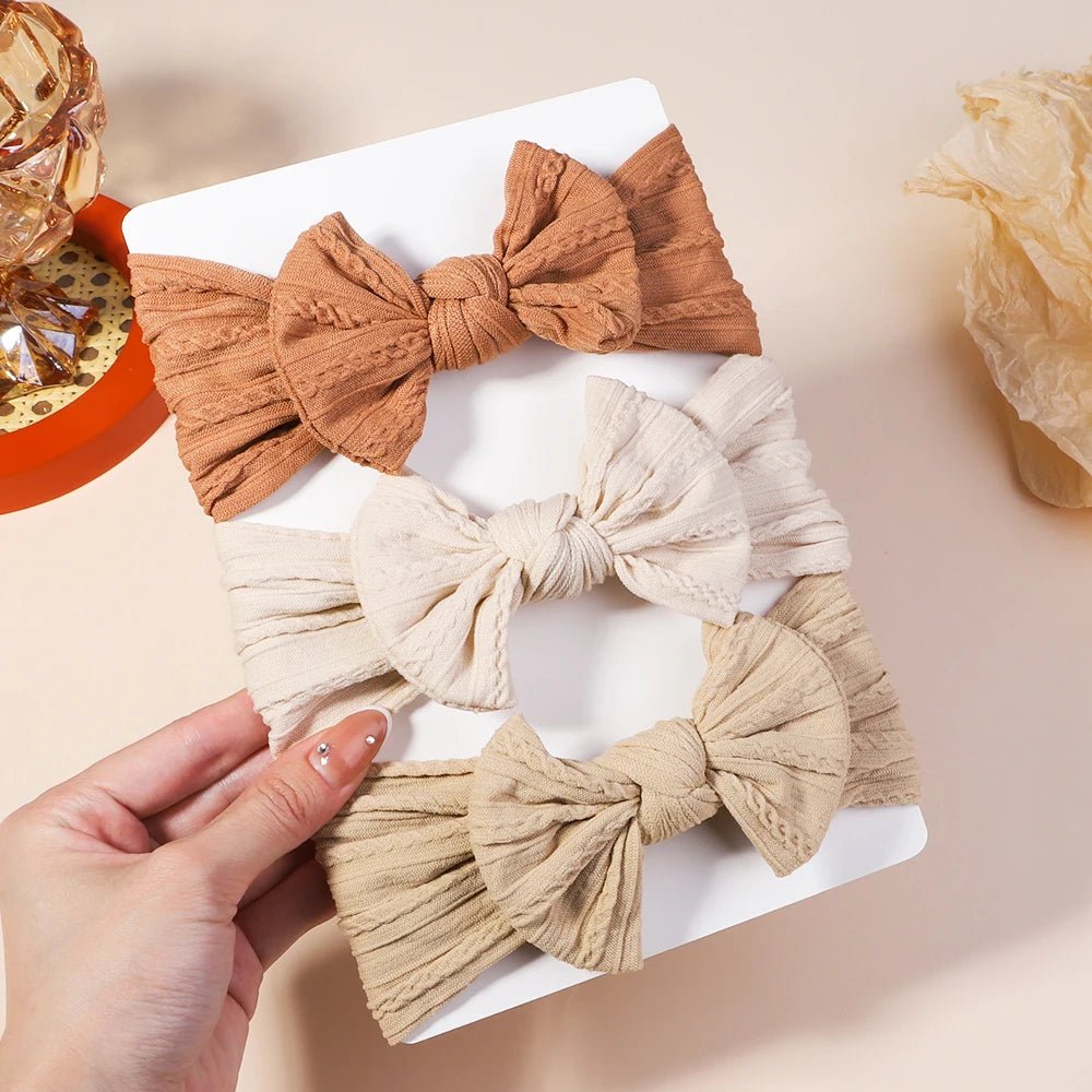 Three fabric hair bows in brown, beige, and cream held by a hand on a light background.