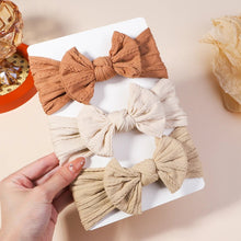 Three fabric hair bows in brown, beige, and cream held by a hand on a light background.