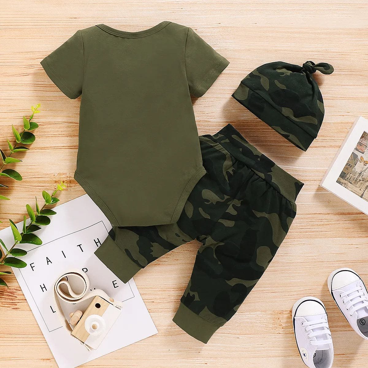 Green baby outfit with matching hat and camouflage pants on a wooden surface.