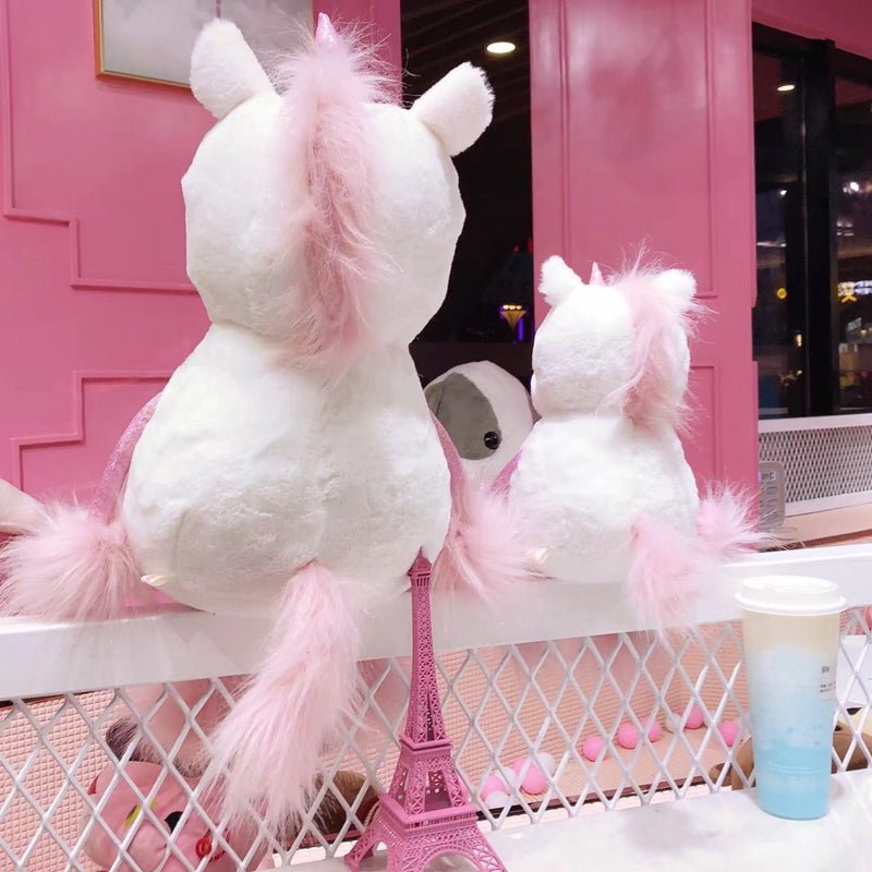 Two white plush unicorns with pink hair on a white table against a pink wall.