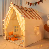 Children's play tent with striped roof and teddy bear inside, decorated with string lights.