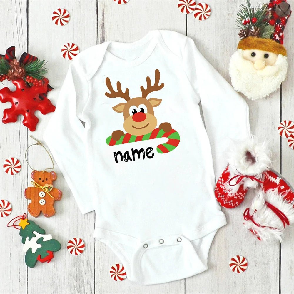 White baby onesie with a reindeer design and candy cane, surrounded by Christmas decorations on a wooden surface. (option 3)