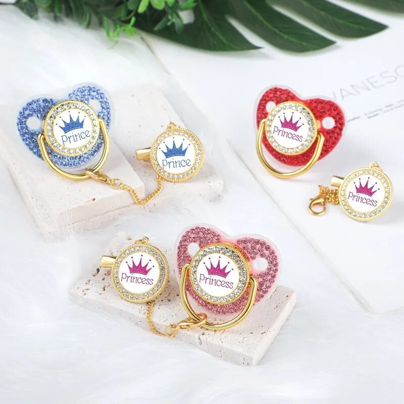 Luxury Crown Pacifier & Clip Set, decorative pacifiers with Prince and Princess text on a white surface with green leaves in the background, for teething relief and calm naps.
