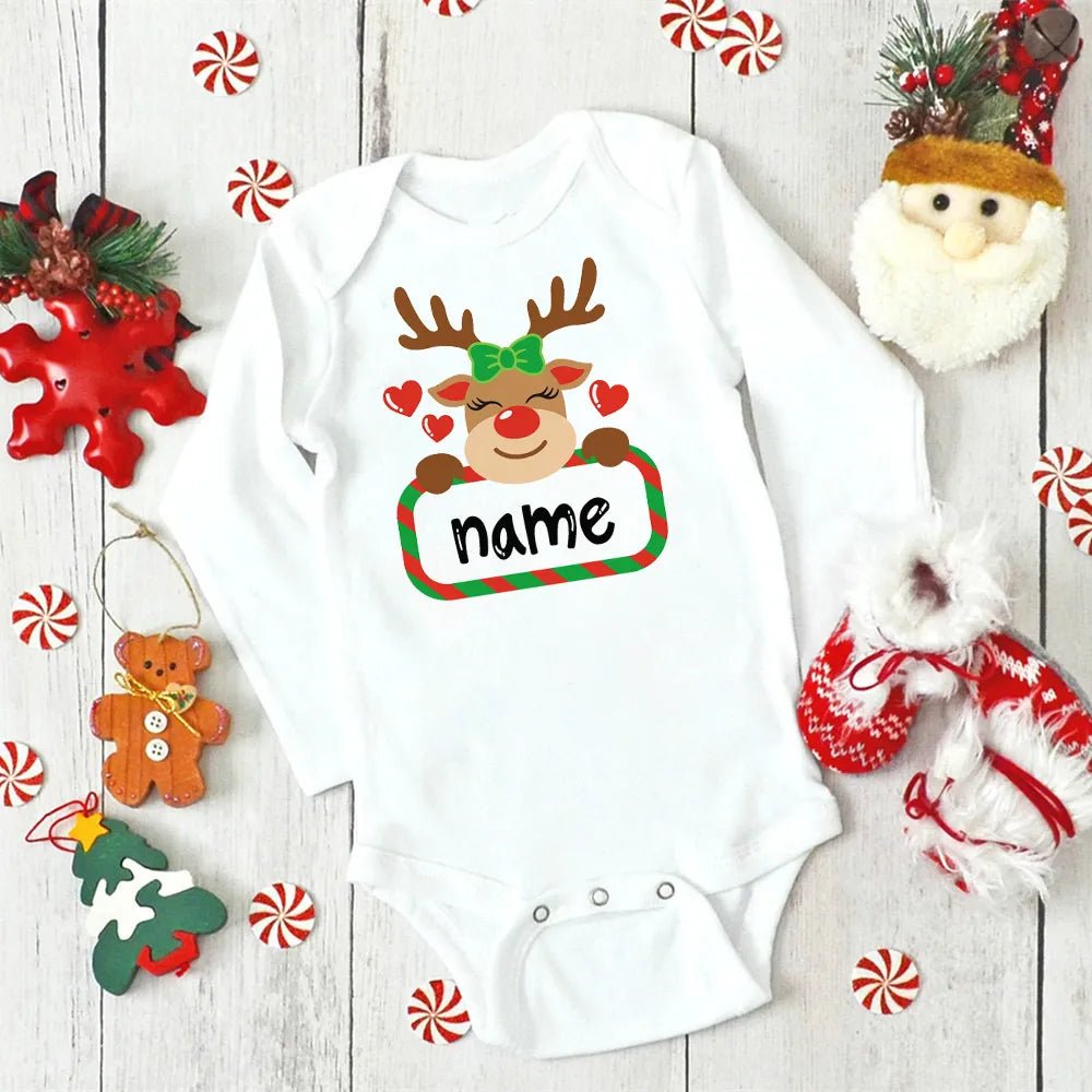 White baby onesie with a reindeer design and 'name' text, surrounded by Christmas decorations on a wooden surface.