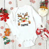 White baby onesie with a reindeer design and 'name' text, surrounded by Christmas decorations on a wooden surface.