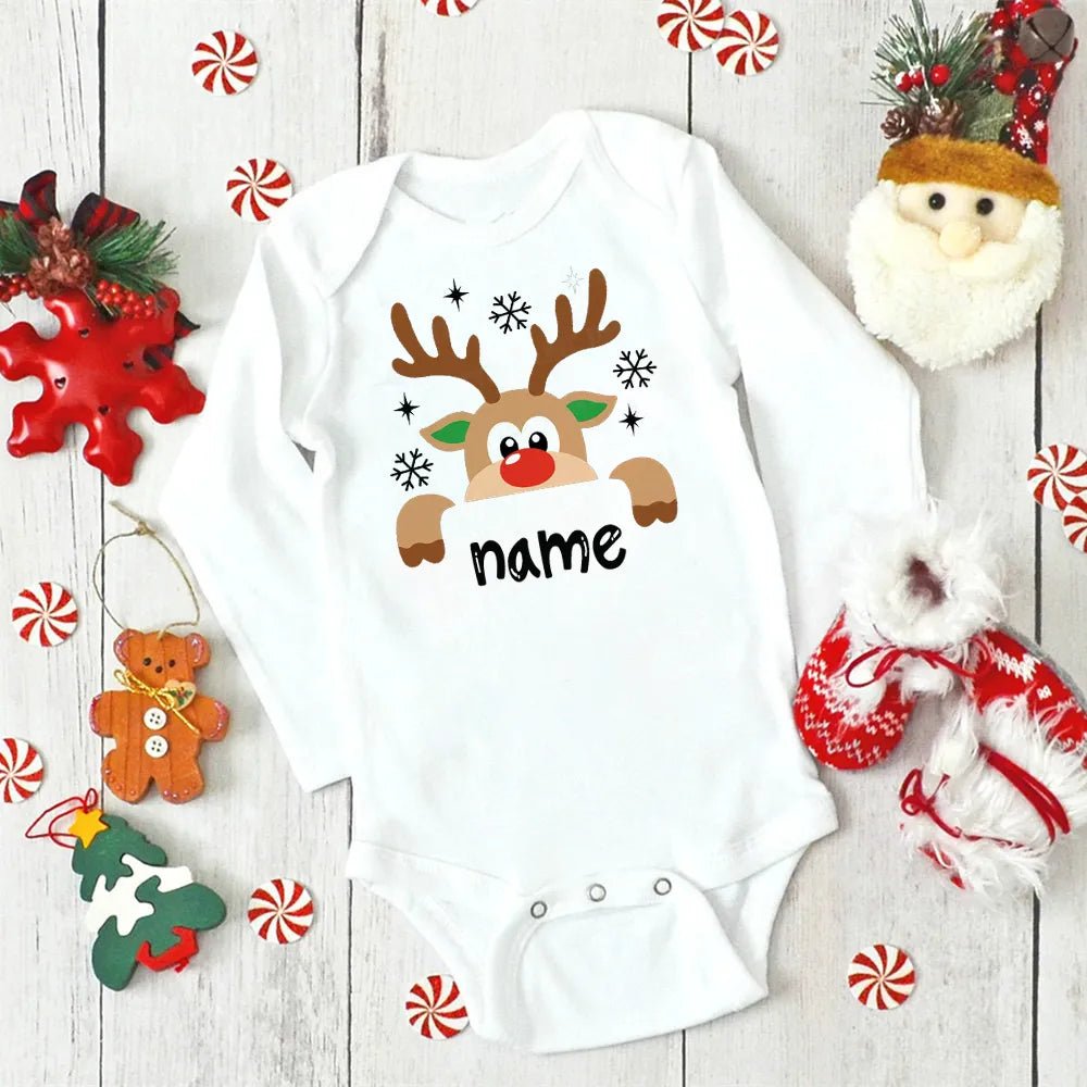 White baby onesie with a reindeer design and 'name' text, surrounded by Christmas decorations on a wooden surface. (option 9)