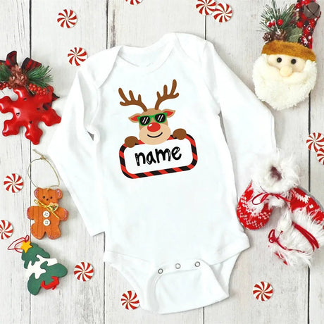 White baby onesie with reindeer design and 'name' text, surrounded by Christmas decorations on a wooden surface.
