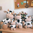 Four cow-themed plush toys on a wooden surface with a bulletin board in the background.