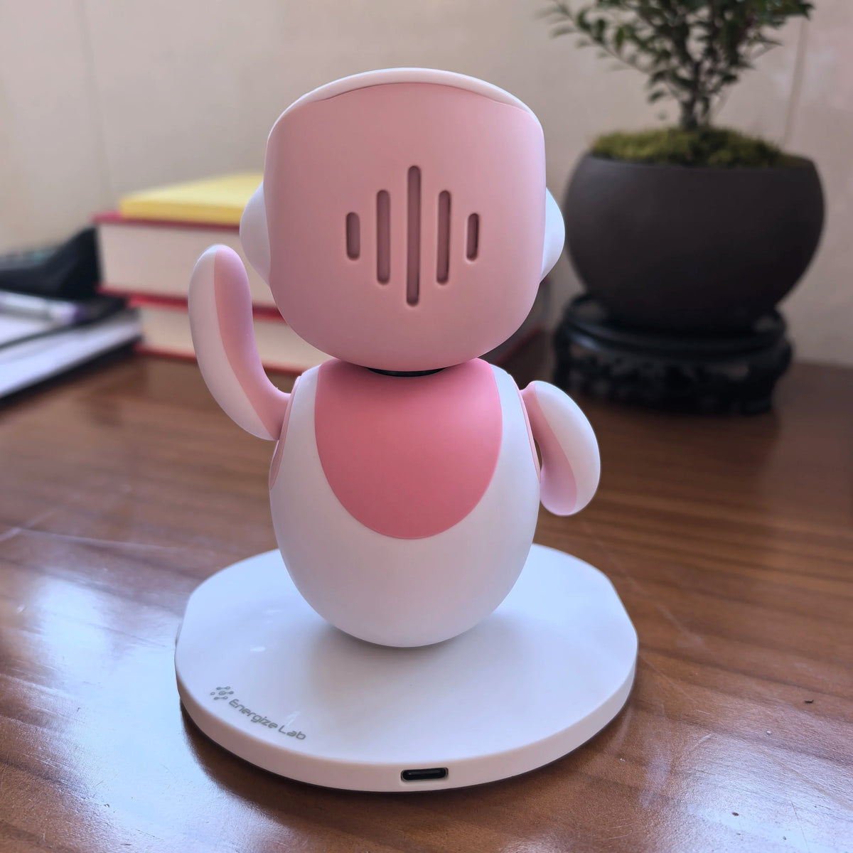 Back of a pink and white desktop robot pet with speaker slots on its head, on a round base on a wooden desk