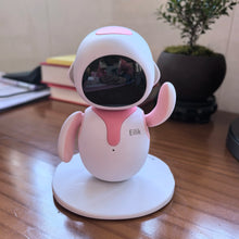 Pink desktop robot pet with a dark screen face and a raised arm on a wooden desk beside a small plant