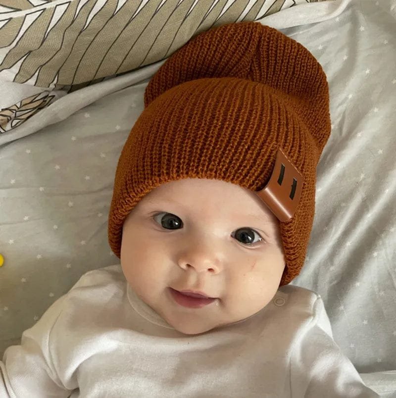 Baby wearing a brown knit hat with a leather strap on a light-colored blanket.