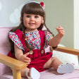 Doll with long brown hair wearing a red dress with floral top and white shoes, sitting on a pink chair.
