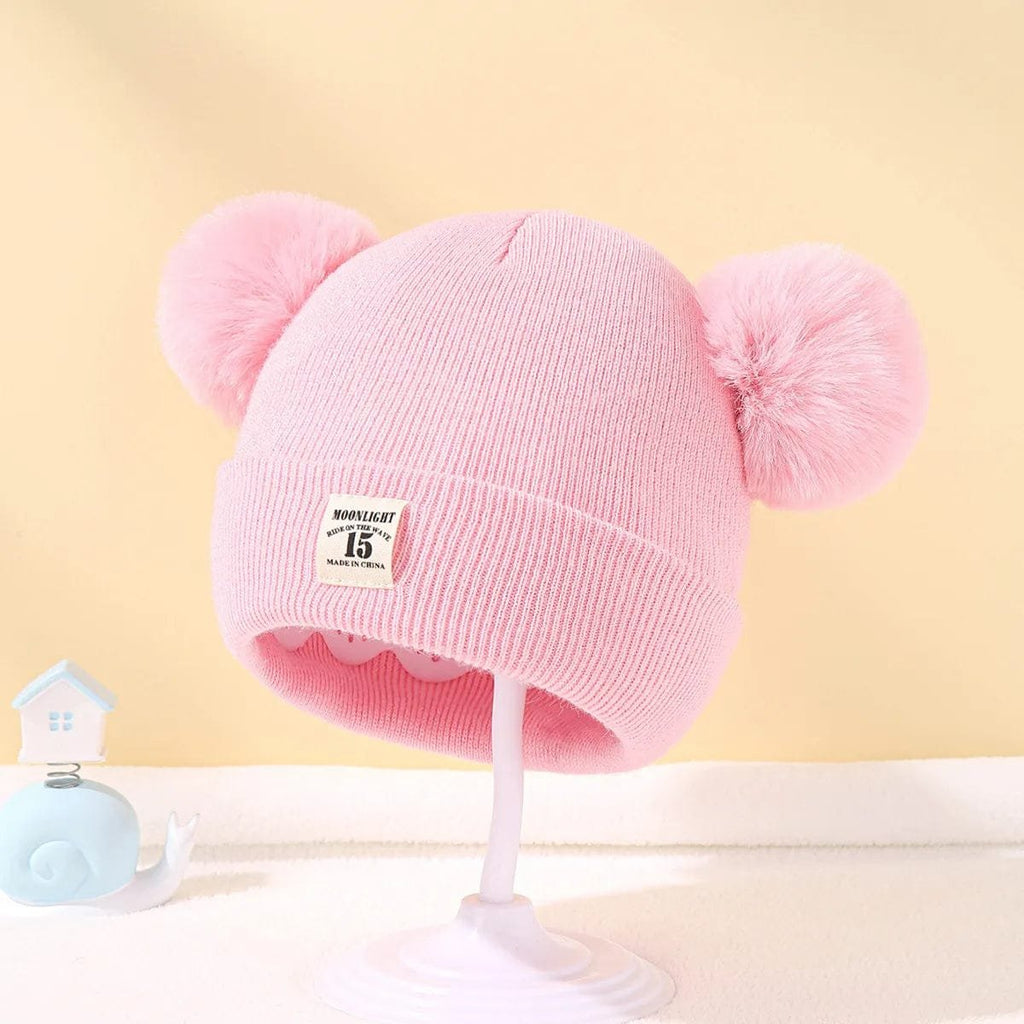 Pink knitted baby hat with two pom-poms on a yellow background