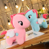 Two plush toys, a pink and a blue unicorn, on a wooden surface with a blurred indoor background.