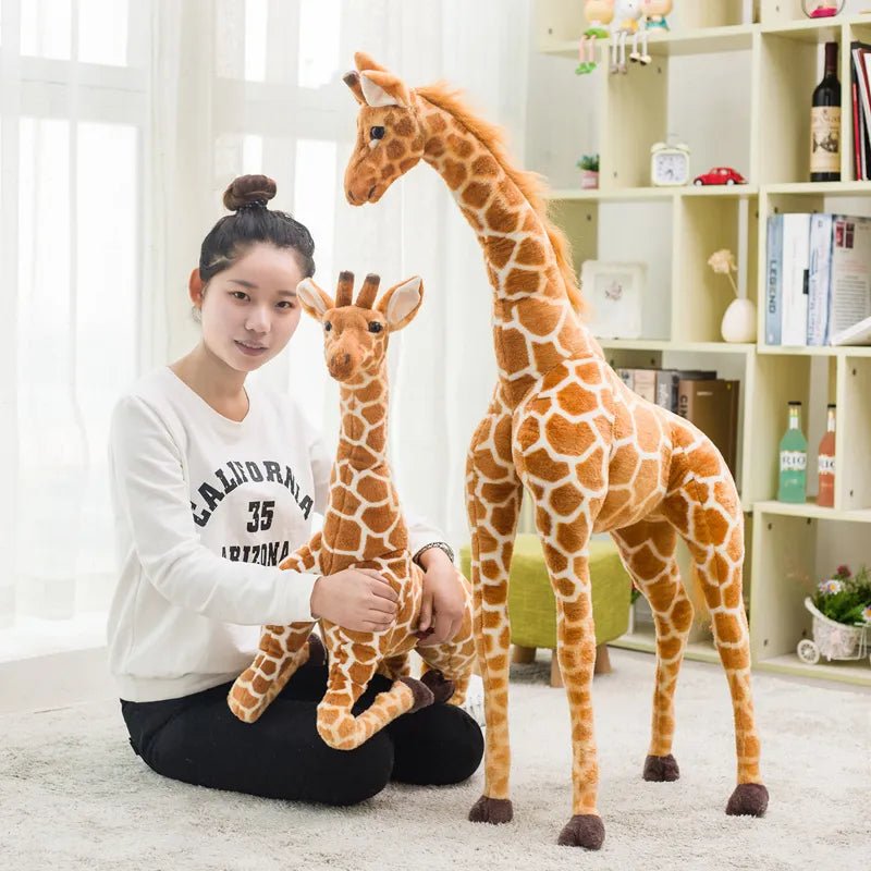 Woman holding a small giraffe plush toy next to a large giraffe plush toy in a room with a bookshelf.