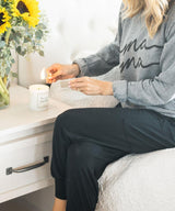 Person sitting on a bed with a gray sweatshirt and black pants, next to a white drawer with a candle and sunflower arrangement.