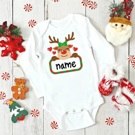 White baby onesie with a reindeer design and 'name' text, surrounded by Christmas decorations on a wooden surface.