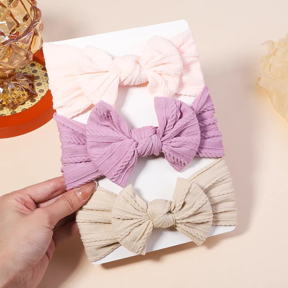 Three fabric hair bows in pink, purple, and beige on a white box with a hand holding one of the bows.