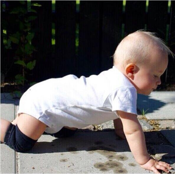 Baby in a white onesie crawling outdoors while wearing knee protectors for safe movement on rough ground.