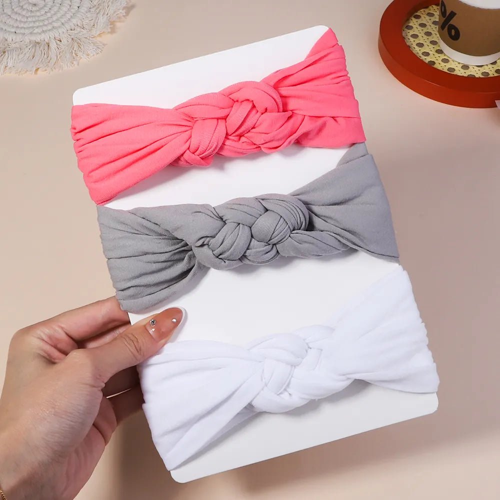 Three headbands in pink, gray, and white on a white surface with a hand holding the white one.