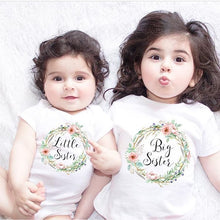 Two children wearing t-shirts with 'Little Sister' and 'Big Sister' text, lying on a white surface. (option 3)