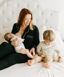 Woman in pajamas holding a baby and sitting on a bed with a toddler.