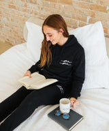 Person sitting on a bed reading a book with a mug and sunglasses beside them.